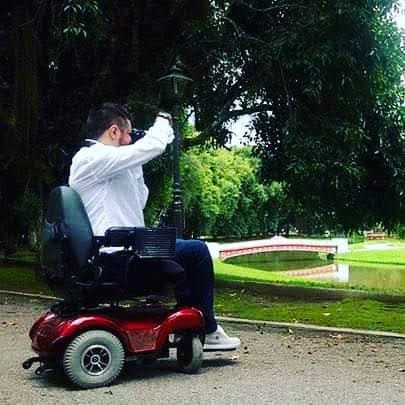 Berguim fotografando em um parque, em sua cadeira de rodas motorizada, com uma ponte ao fundo.